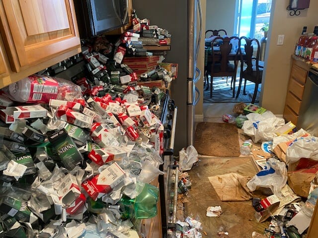 Image shows a kitchen room where the stove and kitchen counters are filled with trash, empty cigarette packs and pizza boxes. There's also trash on the floor. Kitchen stove cannot be used.